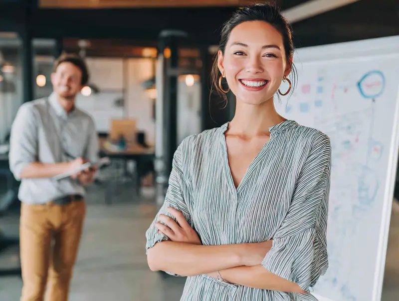 A confident young woman stands in a modern office in front of a whiteboard and symbolizing empowering employees to take ownership and act independently.