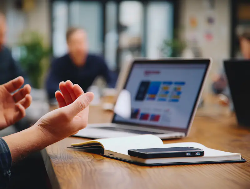 A person gesturing during a meeting, with a laptop and notebook on the table; symbolizing open communication when managing resistance in teams.