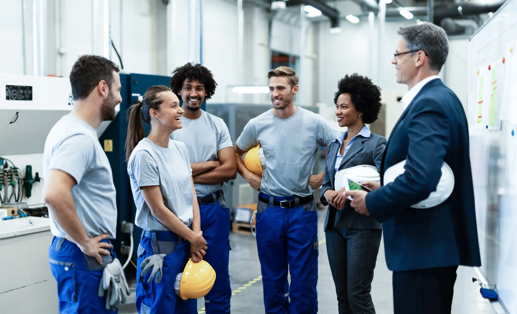 Führungskräfte und Mitarbeitende mit Schutzhelmen im Gespräch in einer Produktionshalle